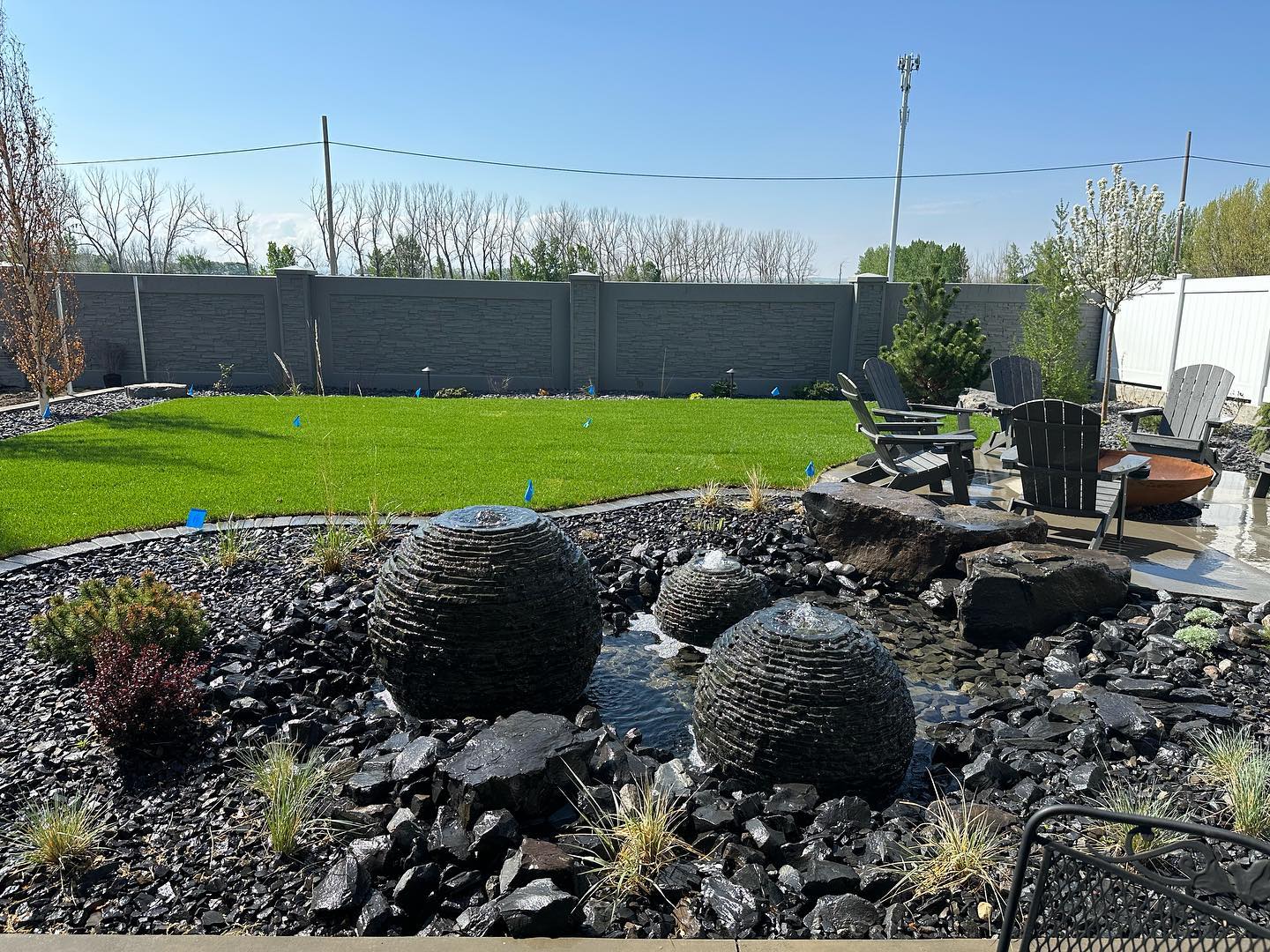 Triple basalt sphere water feature in black rock bed with fresh sod lawn, Adirondack fire pit lounge, and stone fence behind