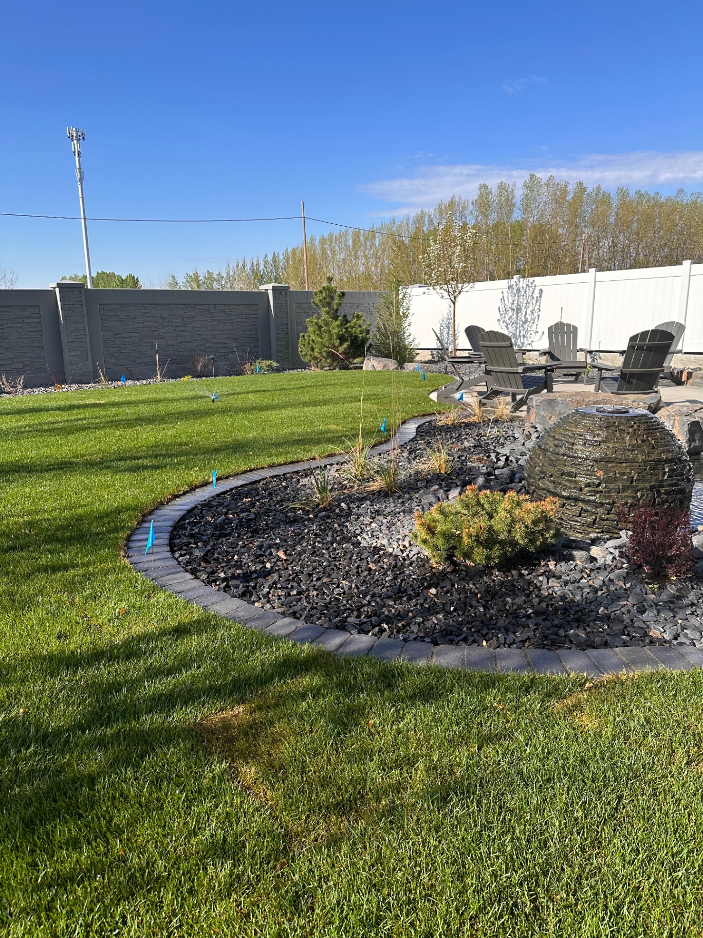 Backyard with curved paver-edged rock bed, basalt sphere water feature, and Adirondack chairs around a fire pit