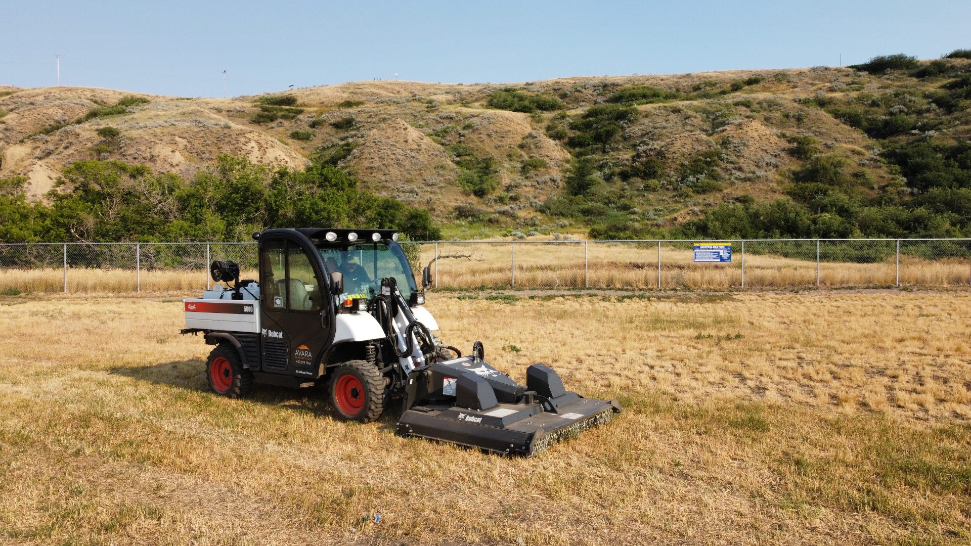 Avara Property Care Bobcat Toolcat with rotary brush cutter mowing dry grass on an industrial site with coulee hills in the background