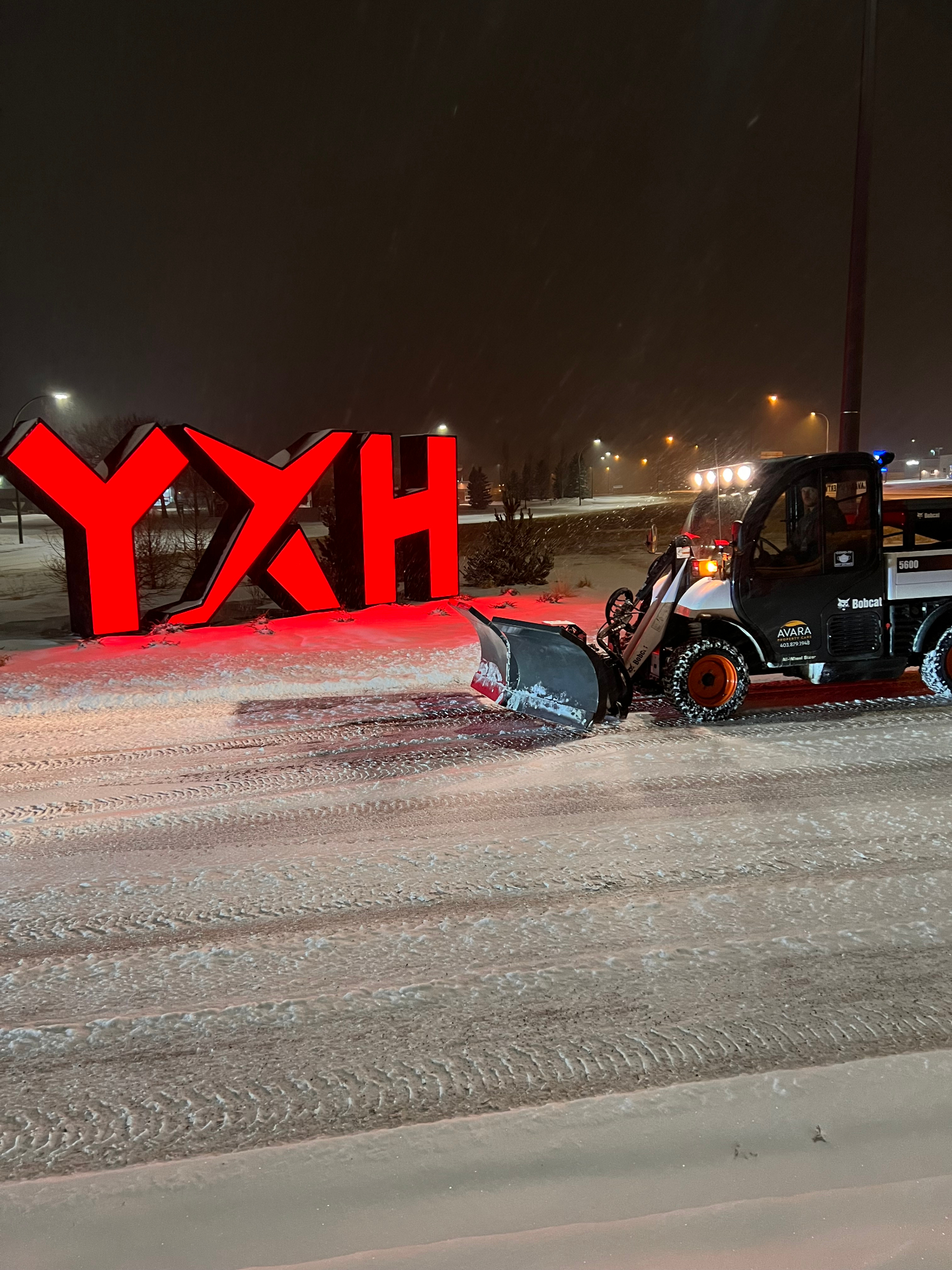 Avara Property Care Bobcat 5600 Toolcat with snow plow clearing the YXH Medicine Hat Airport entrance during a winter storm at night