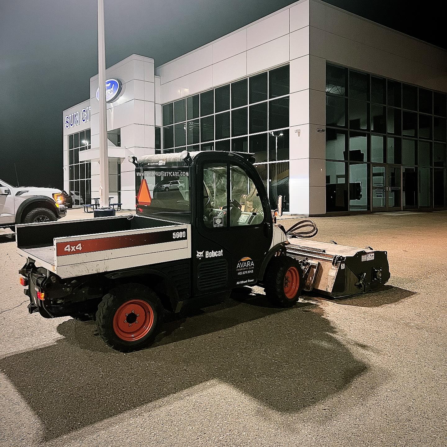 Avara Property Care Bobcat Toolcat with sweeper attachment servicing a commercial dealership parking lot at night