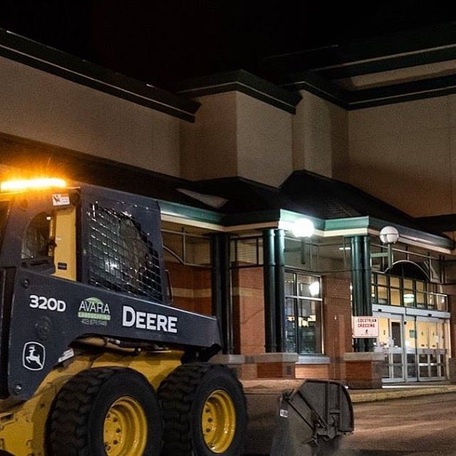 Avara John Deere 320D skid steer servicing the entrance of a retail center at night